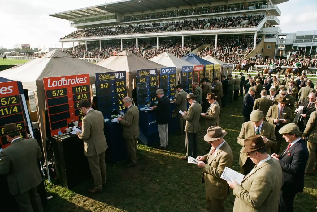 Wide view of a busy UK racecourse betting ring with on-course bookmakers and racegoers studying the odds boards